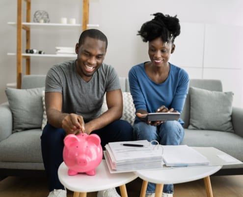 Couple sitting on a couch reviewing finances, placing money into a pink piggy bank with bills and paperwork on the table.