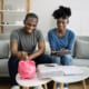 Couple sitting on a couch reviewing finances, placing money into a pink piggy bank with bills and paperwork on the table.