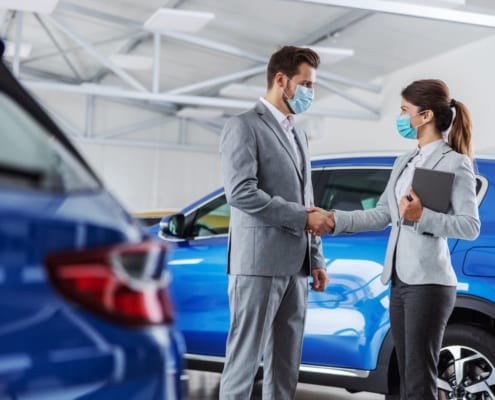 Man and woman wearing face masks shaking hands in a car showroom, standing beside a blue vehicle.
