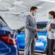 Man and woman wearing face masks shaking hands in a car showroom, standing beside a blue vehicle.