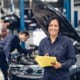 a lady car mechanic smiling at the camera with a clipboard in her hands with a man mechanic inspecting an engine