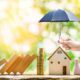 Umbrella protecting model house family figures and coins symbolizing home insurance coverage