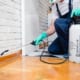 Pest control worker wearing gloves and a mask, kneeling on a hardwood floor while spraying pesticide along the base of a wall with a handheld sprayer.