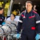 an EMT looking at the camera while another emt helps a patient on a stretcher into an ambulance