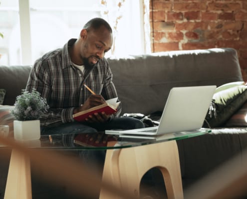 man writing in his notebook with a laptop on a table infront of him