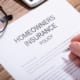 Close-up of a homeowners insurance policy document on a wooden desk, with a hand holding a pen ready to sign and eyeglasses placed beside the paperwork.