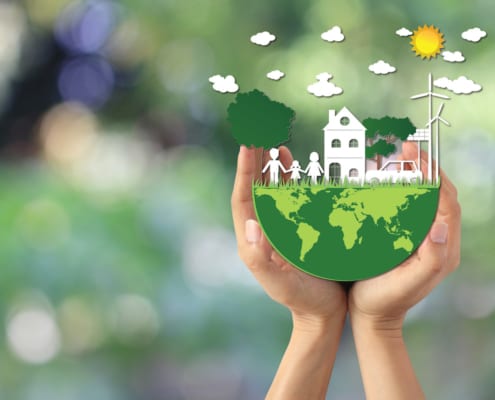 Hands holding a small globe with paper-style house, family, trees, car, wind turbine, and sun, symbolizing environmental protection and sustainable living.
