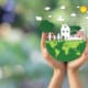 Hands holding a small globe with paper-style house, family, trees, car, wind turbine, and sun, symbolizing environmental protection and sustainable living.