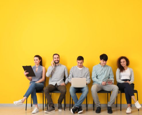 Group of job applicants sitting in a row using laptop, tablet, phone, and clipboard against yellow wall