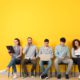 Group of job applicants sitting in a row using laptop, tablet, phone, and clipboard against yellow wall