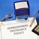 Homeowners insurance policy document on a desk with a calculator, pen, eyeglasses, and a stamp.