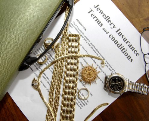 Gold jewelry and watch placed on jewellery insurance policy document with glasses and folder on desk