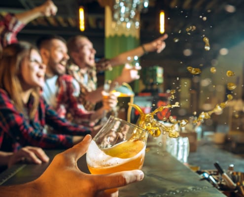 Hand holding a glass of beer mid-splash at a bar while a group of friends in the background cheer and celebrate watching a game on TV.