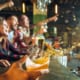 Hand holding a glass of beer mid-splash at a bar while a group of friends in the background cheer and celebrate watching a game on TV.