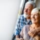 Happy senior couple standing by window, embracing and smiling.