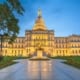 Illuminated state capitol building with a large dome at dusk, viewed from the front across a walkway and lawn, with a statue centered in front and trees framing the scene.
