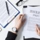Person reviewing an insurance policy document on a desk, with a clipboard contract, pen, eyeglasses, tablet, and wristwatch visible