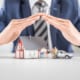 Hands protecting miniature house, family figures, and car on table