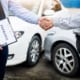 Two men shaking hands in front of damaged cars, one holding a clipboard labeled “Insurance”