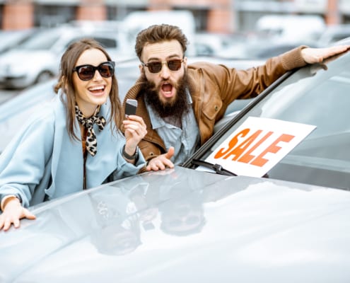 Excited couple holding car key next to vehicle with “Sale” sign in windshield at dealership