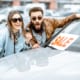 Excited couple holding car key next to vehicle with “Sale” sign in windshield at dealership