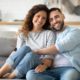 Smiling couple sitting together on couch at home with arms around each other