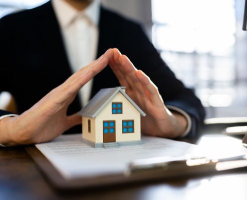 home insurance agent with papers Infront of him with a house figure on them with his hands over the house making a roof
