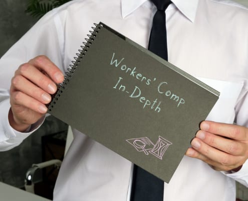 Business professional holding a notebook labeled “Workers’ Comp In-Depth” while wearing a white shirt and black tie